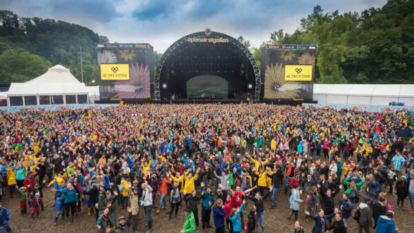 A large outdoor crowd at a music festival with people wearing colorful raincoats, standing on muddy ground, cheering in front of a stage with the name 'Passenger' displayed and banners reading 'Openair St.Gallen'.