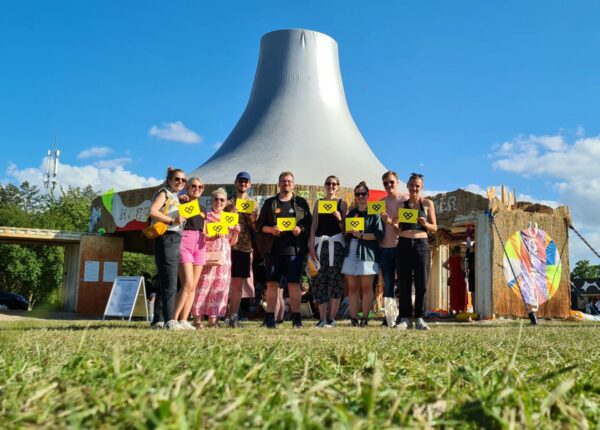 A group of people standing outdoors holding signs with heart and exclamation mark symbols in front of a large funnel-shaped structure on a sunny day.