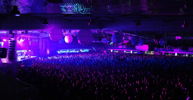 A large indoor concert venue filled with a crowd under blue lighting, with visible stage and hanging spherical decorations.