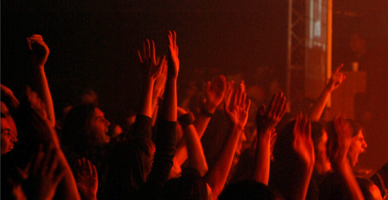 Crowd of people at a concert with raised hands, illuminated by red stage lighting.