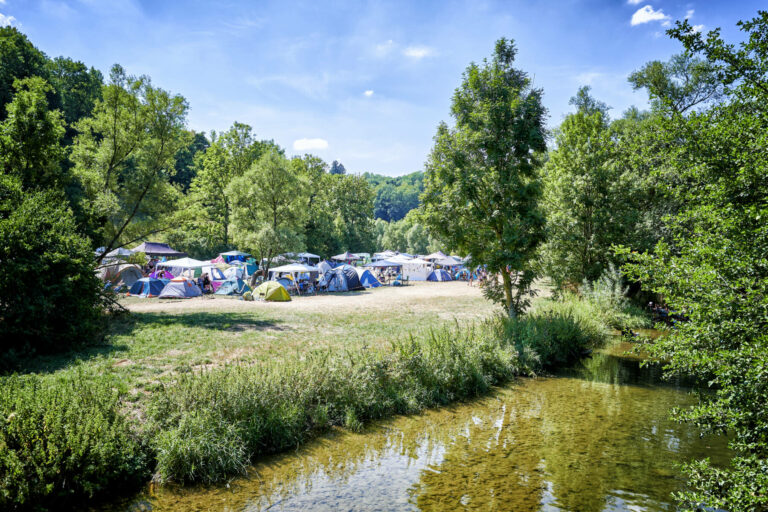 A scenic outdoor camping area with a collection of tents set up on the grassy bank of a calm river surrounded by lush green trees under a clear blue sky.