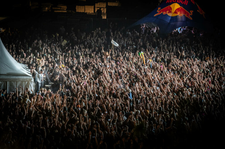 An enthusiastic crowd at a nighttime event, with many hands raised in the air under the glow of stage lighting, and a prominent Red Bull tent in the background.