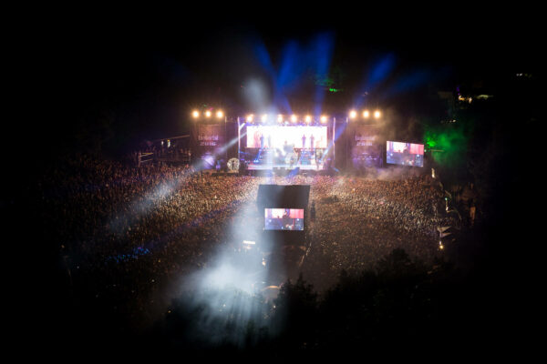 A large outdoor night concert with a dense crowd watching a lit stage, enhanced by beams of blue light and a screen showing performers, with trees surrounding the area.