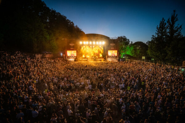 A large outdoor concert at dusk with a crowd of people facing a lit stage, surrounded by trees.