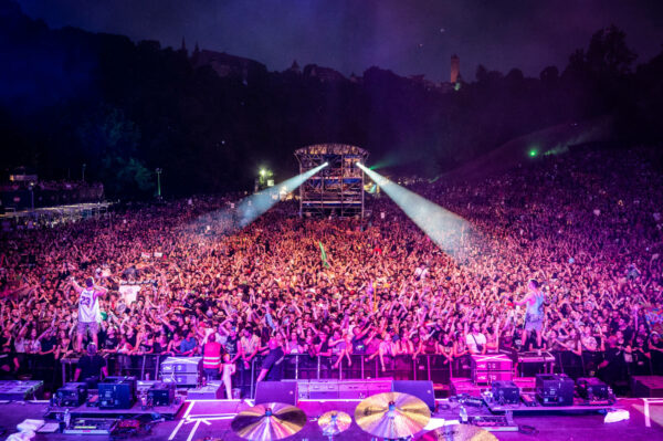 A vibrant outdoor concert scene at night with a large crowd of fans, stage lighting, and musicians performing, surrounded by trees and a hill in the background.