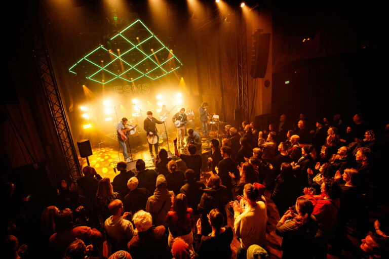 The Mary Wallopers performing onstage with the audience clapping in a warmly lit concert venue with a neon geometric shape overhead and the letters 