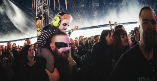 A man with face paint wearing sunglasses and a beard carries a child with similar face paint and protective headphones on his shoulders, both enjoying themselves amidst a crowd at what appears to be a music festival.