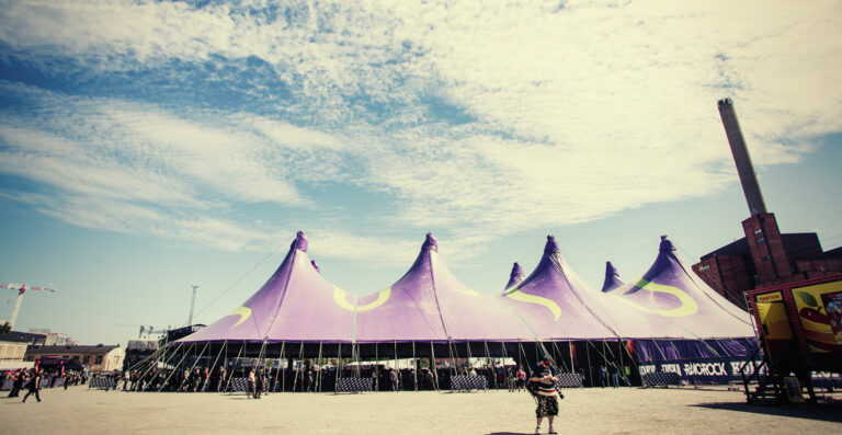 A large purple and white circus tent set up in an open area with clear skies and a few clouds above, industrial buildings in the background, and a few individuals walking in the foreground.