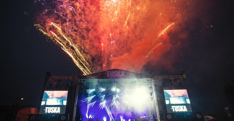 A vibrant firework display lights up the night sky above an outdoor music festival stage with the sign 