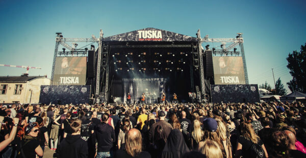 A large crowd of people at an outdoor music festival with the band Apocalyptica performing on stage under a banner celebrating the Tuska festival's 20th anniversary.