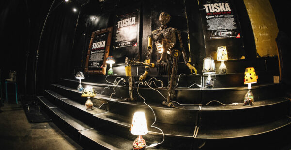 A dimly lit stage with a skeletal metal sculpture holding a guitar, surrounded by various unique lamps on each stair level and concert posters on the wall.