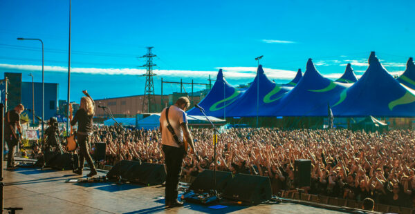 A band performing on an outdoor stage in front of a cheerful crowd with hands raised under a clear blue sky, with event tents in the background.