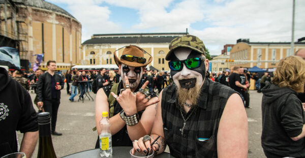 Two individuals with face paint and metal-inspired attire posing for a photo, with one making a rock hand gesture, amidst a crowd at what appears to be a music festival.