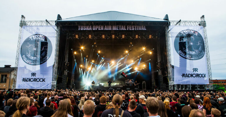 Crowd at an outdoor metal festival with a stage set and large screens displaying event branding on either side.
