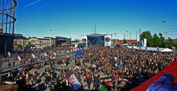 Outdoor music festival crowd with a large stage in the background and a clear blue sky above.
