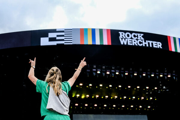 A person from behind with raised arms making the sign of the horns gesture in front of the Rock Werchter stage with the festival's name displayed.