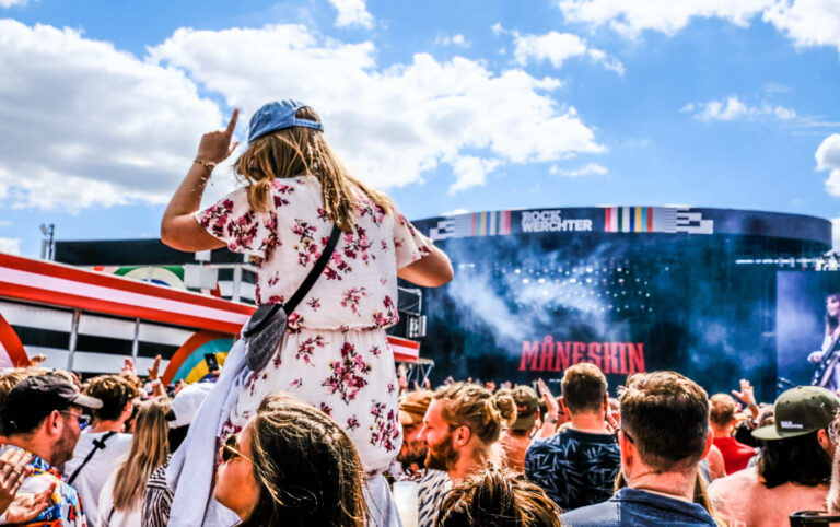 A crowd of people enjoying a live concert at the Rock Werchter festival with a view of the stage displaying the band name Måneskin under a bright sky. A person in a floral dress is prominently raising their hand near the center.