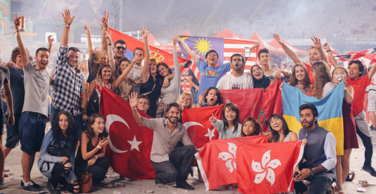 A large group of smiling people, some standing and some squatting, holding various national flags including Turkey, Malaysia, and Romania, at a crowded event with a cheerful atmosphere.