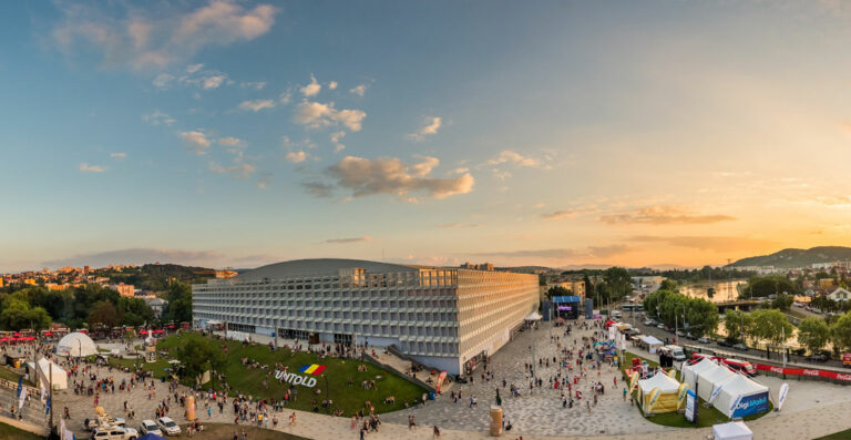 Panoramic view of a festival scene at dusk, with crowds of people in an open square next to a modern building, promotional tents and stands around, overlooking a calm river and hills in the distance under a clear sky with some clouds.