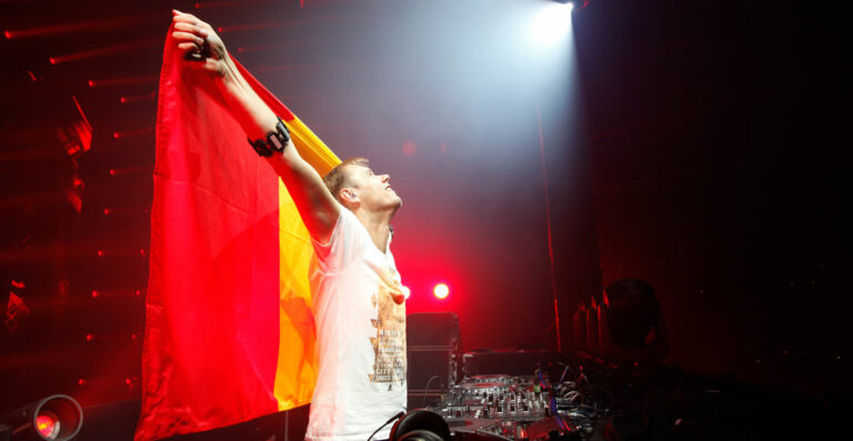 A DJ on stage raising a flag with his eyes closed, bathed in dramatic stage lighting with DJ equipment in the foreground.