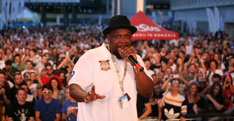 A man in a white shirt and black hat performing on a stage with a microphone in hand, addressing a large outdoor audience.
