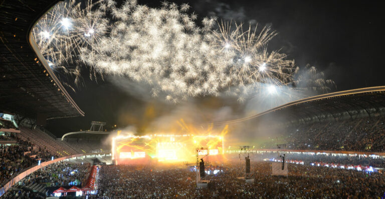 A large crowd at a nighttime stadium event with a spectacular fireworks display overhead.