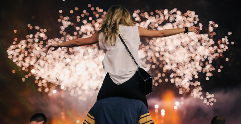 A woman with outstretched arms standing in front of a sparkling fireworks display at night.