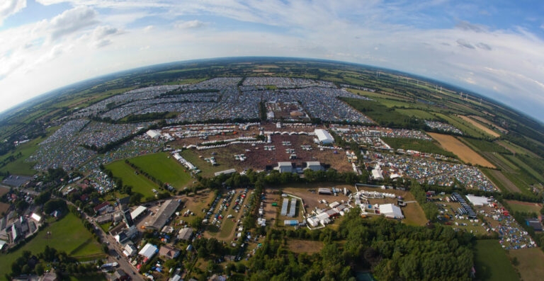 Aerial view of a large outdoor festival with numerous tents and structures, surrounded by green fields and trees, under a clear sky.