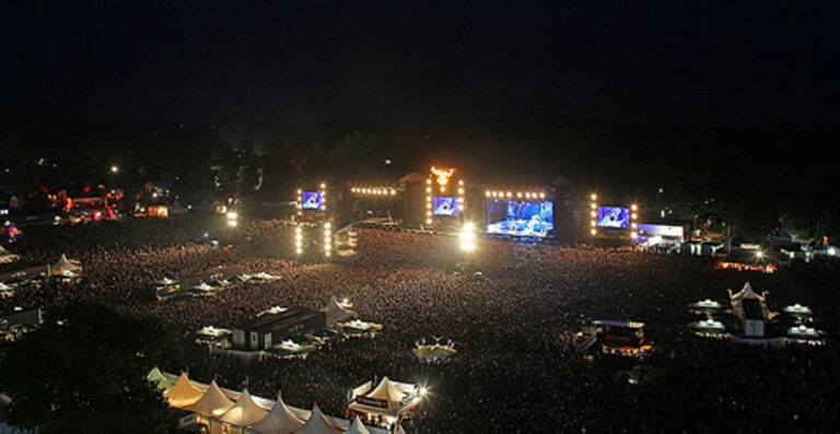 A nighttime view of a crowded outdoor music festival with a brightly lit stage and surrounding festival area.