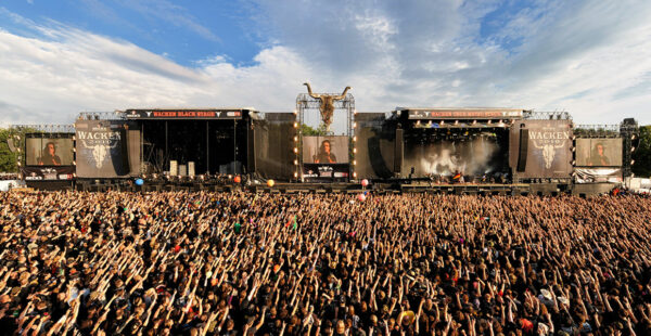 A large crowd of fans at the Wacken Open Air festival cheering in front of a stage with live band performance, large screens, and festival banners under a clear sky.