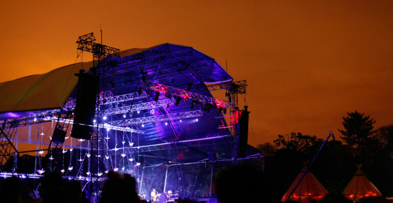 Outdoor concert stage at night with vibrant purple and blue stage lights, silhouettes of the audience in the foreground, and a vivid orange sky in the background.