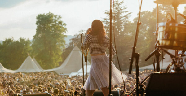 A female performer is standing on stage facing a large audience at an outdoor event, with her back to the camera and a microphone stand in front of her. The setting sun casts a warm glow over the scene.