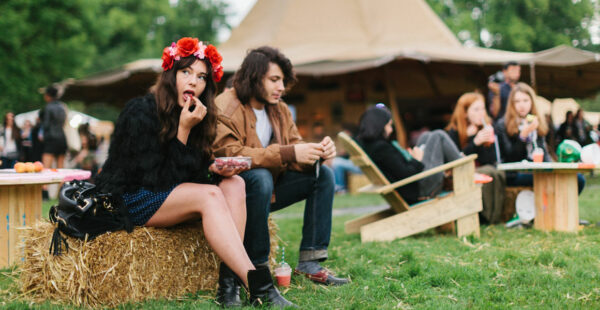 A woman with a floral headband and black clothing eats strawberries while sitting on a hay bale next to a man, with other festival-goers in the background.