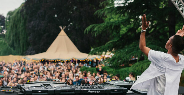 DJ performing at an outdoor music festival with a crowd in front of the stage and a teepee structure in the background.