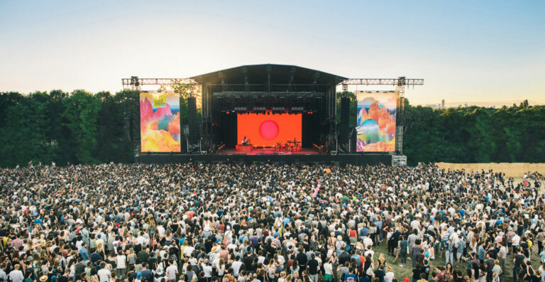 Large outdoor concert with a dense crowd of spectators facing a stage with colorful screens against a backdrop of trees and a clear sky at sunset.