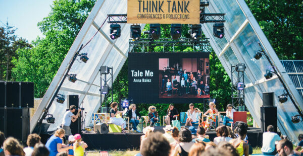 An outdoor conference event at a festival with speakers seated on stage in front of a large audience, under a banner reading 