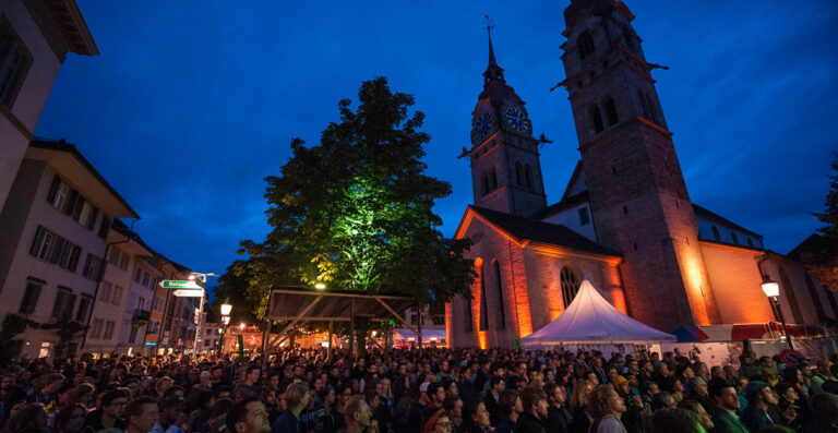 A crowded evening event in a town square with people gathered near a church with illuminated clock towers.