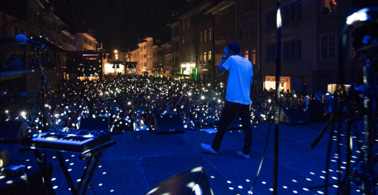 A performer on stage facing a large crowd with raised mobile phones creating a sea of lights at an outdoor nighttime concert.