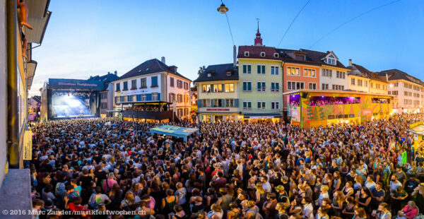 A crowded outdoor music festival at dusk with a large audience gathered in front of a stage in a town square, surrounded by multicolored buildings.