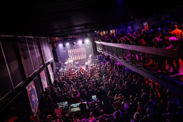 A vibrant indoor concert scene with a crowded audience watching a live band perform on stage, with bright stage lights casting a purple hue over the venue.