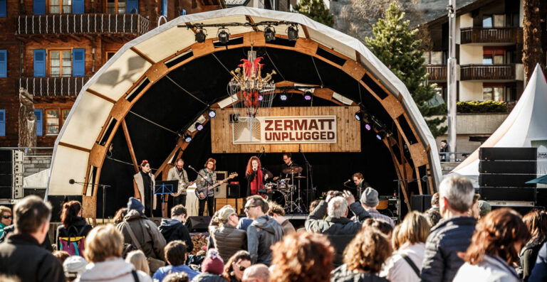 A band performs on an outdoor stage with the sign "ZERMATT UNPLUGGED" at a music event, with a crowd of people watching in the foreground and traditional chalet-style buildings in the background.