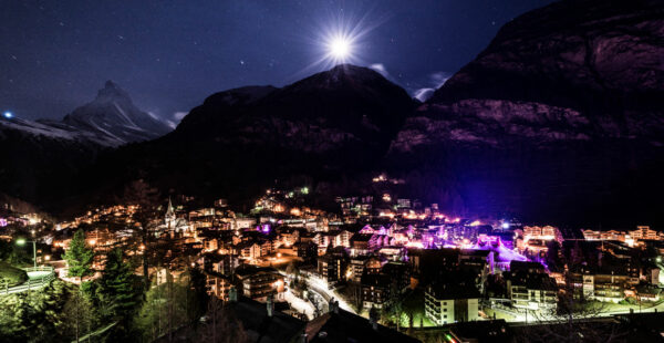 A night view of a lit-up mountain village with the iconic Matterhorn peak in the background under a starry sky with a bright moon.