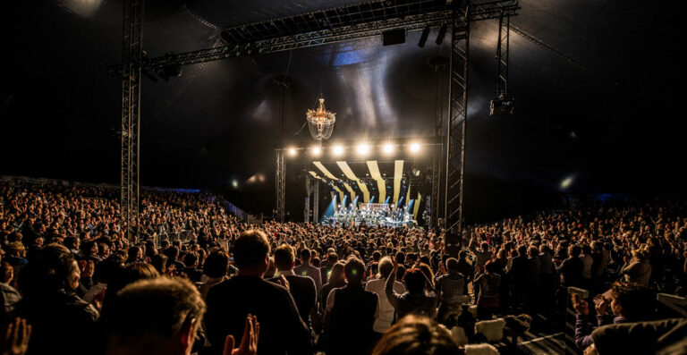 Crowd of spectators at a concert enjoying a live performance inside a large tent with lighting and a chandelier overhead.