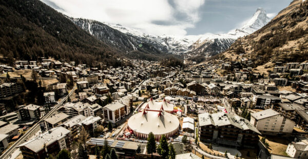 Aerial view of a dense alpine town with traditional buildings, featuring a large circus tent in the foreground and a snow-capped mountain peak in the background.
