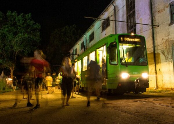 A green tram stopped at night with its doors open as blurred figures of passengers board and alight, next to an old building with illuminated windows.