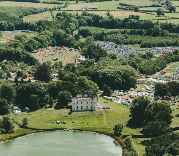 Aerial view of a bustling outdoor festival with tents and attendees near a historic building, surrounded by verdant countryside and a small pond in the foreground.