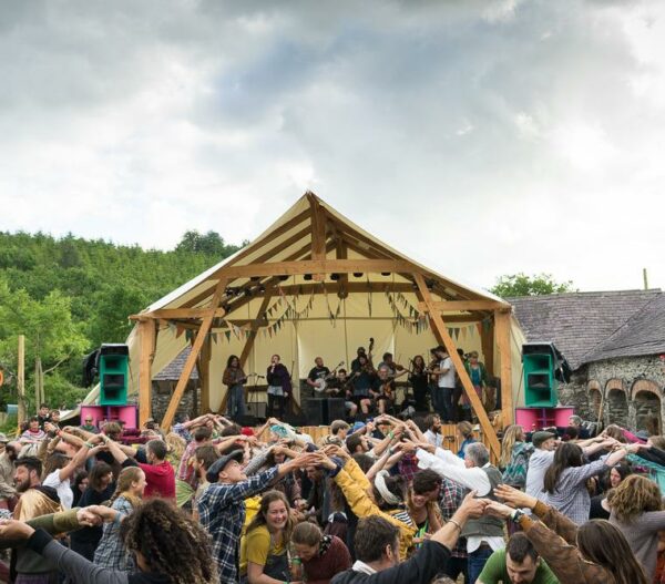 A lively outdoor festival scene with a crowd forming a circle and holding hands high in the air in front of a wooden stage where a band performs, with green hills and a cloudy sky in the background.