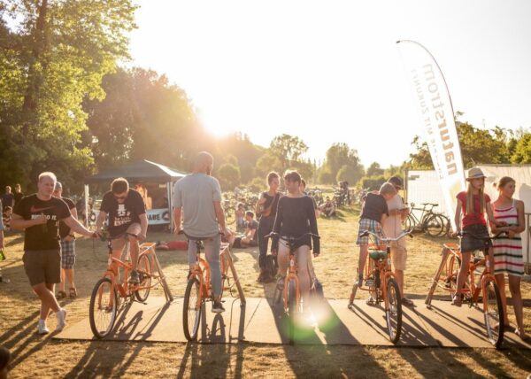 A group of people enjoying a sunny outdoor event with several of them standing and walking with orange bicycles. A promotional white banner is visible on the right, trees in the background, and the sun is low in the sky, suggesting evening time.
