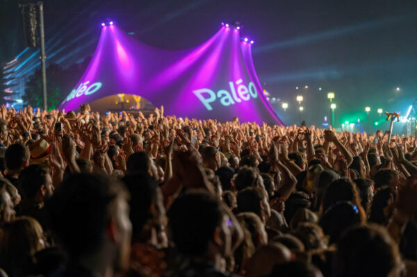 A large crowd of people at a music festival raising their hands in the air with stage lights shining above and the word 