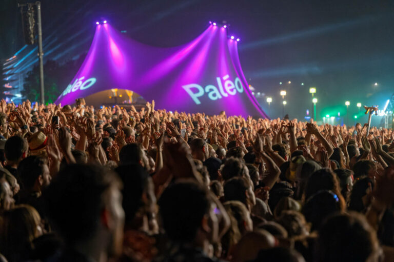 A large crowd of people at a music festival raising their hands in the air with stage lights shining above and the word "Paléo" displayed prominently in the background.